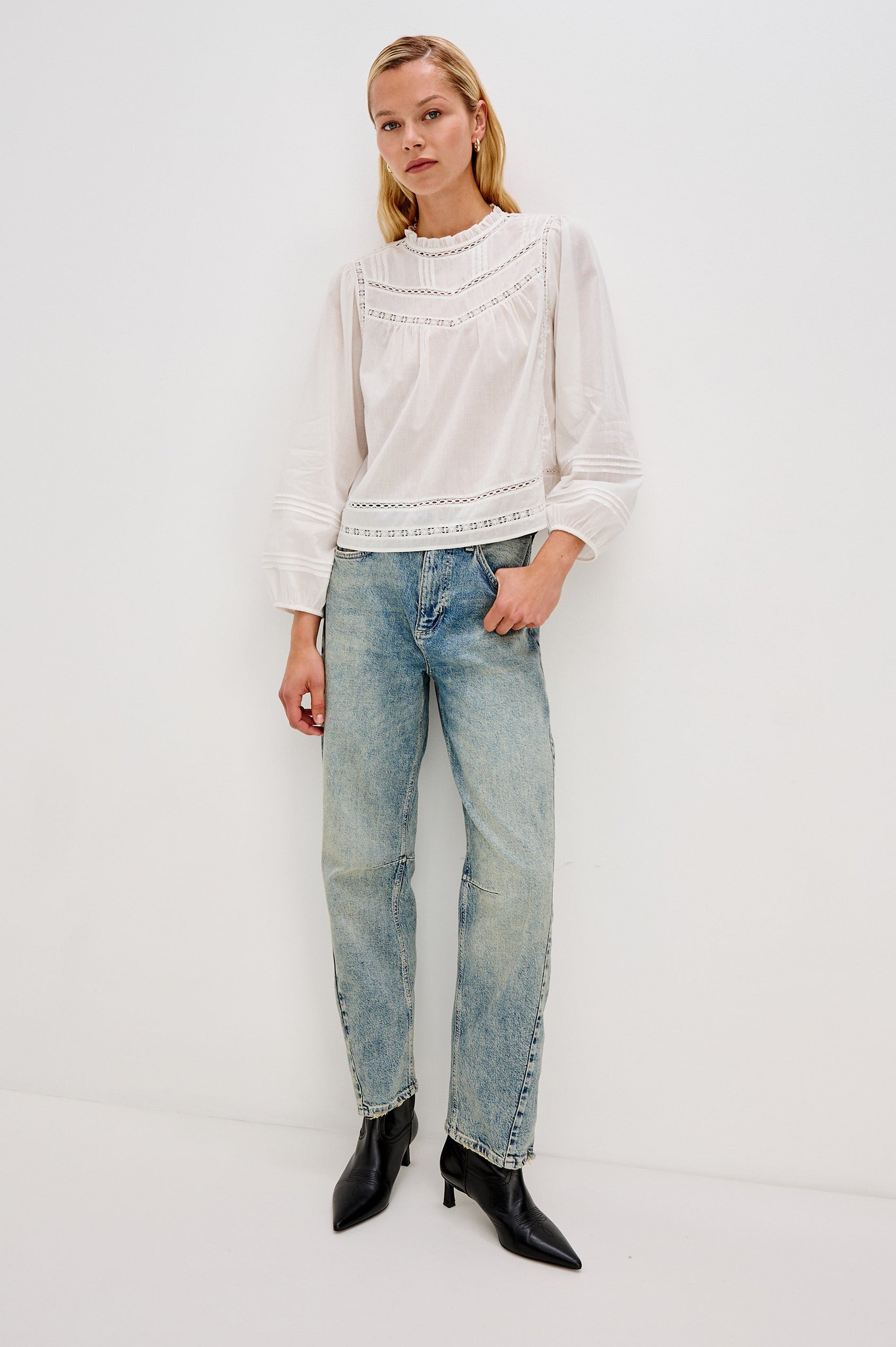 Full body front view of female model wearing our Reyna Top in white. This is a long sleeve crewneck baby doll blouse, with ruffles at the collar. 