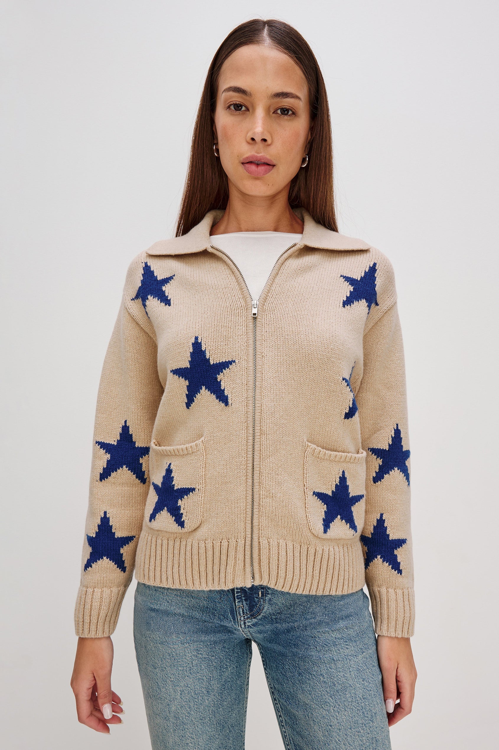 Rhea, a longline camel cardigan in a wool blend, featuring cobalt blue star motifs. The cardigan has a relaxed fit, zip-front closure, and front patch pockets. Front View Featured on Model, Zipped Up