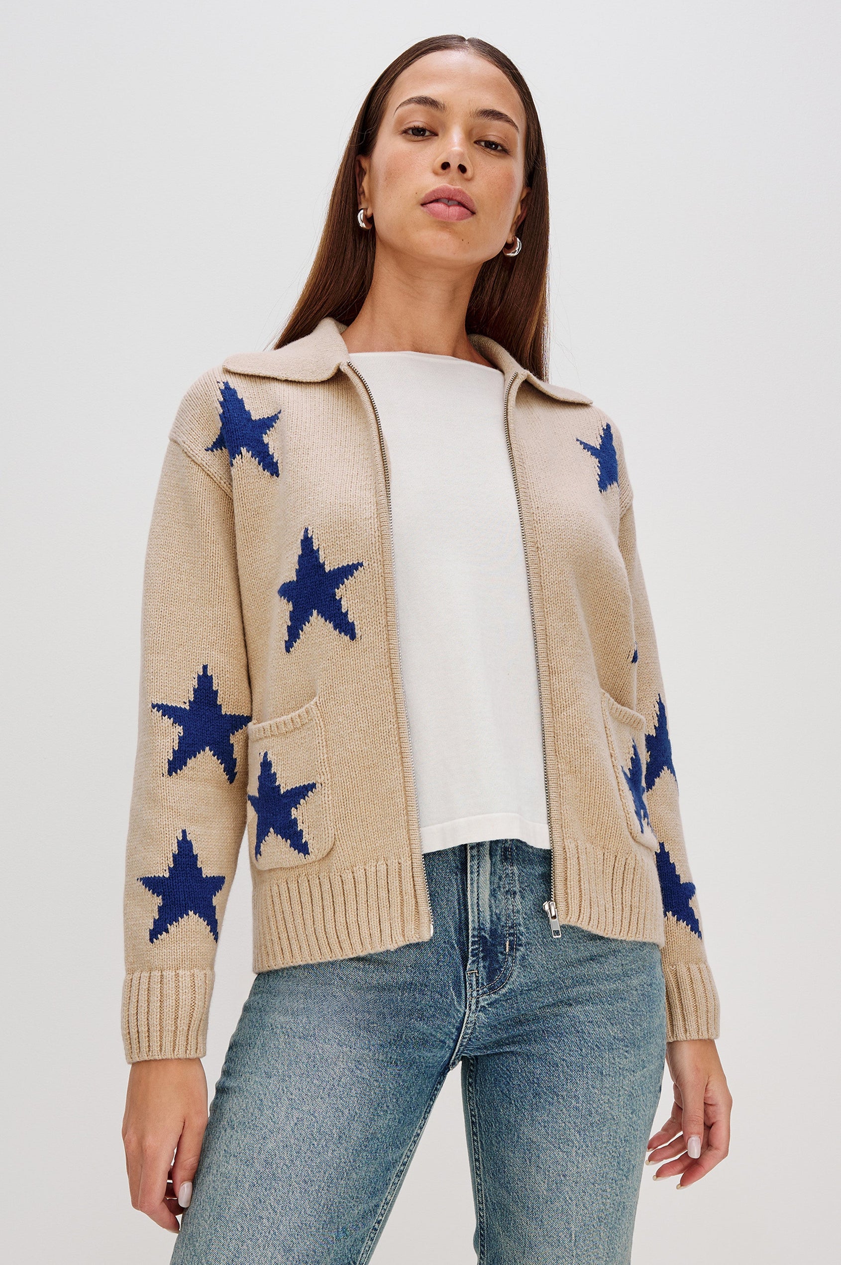 Rhea, a longline camel cardigan in a wool blend, featuring cobalt blue star motifs. The cardigan has a relaxed fit, zip-front closure, and front patch pockets. Front View Featured on Model, Unzipped
