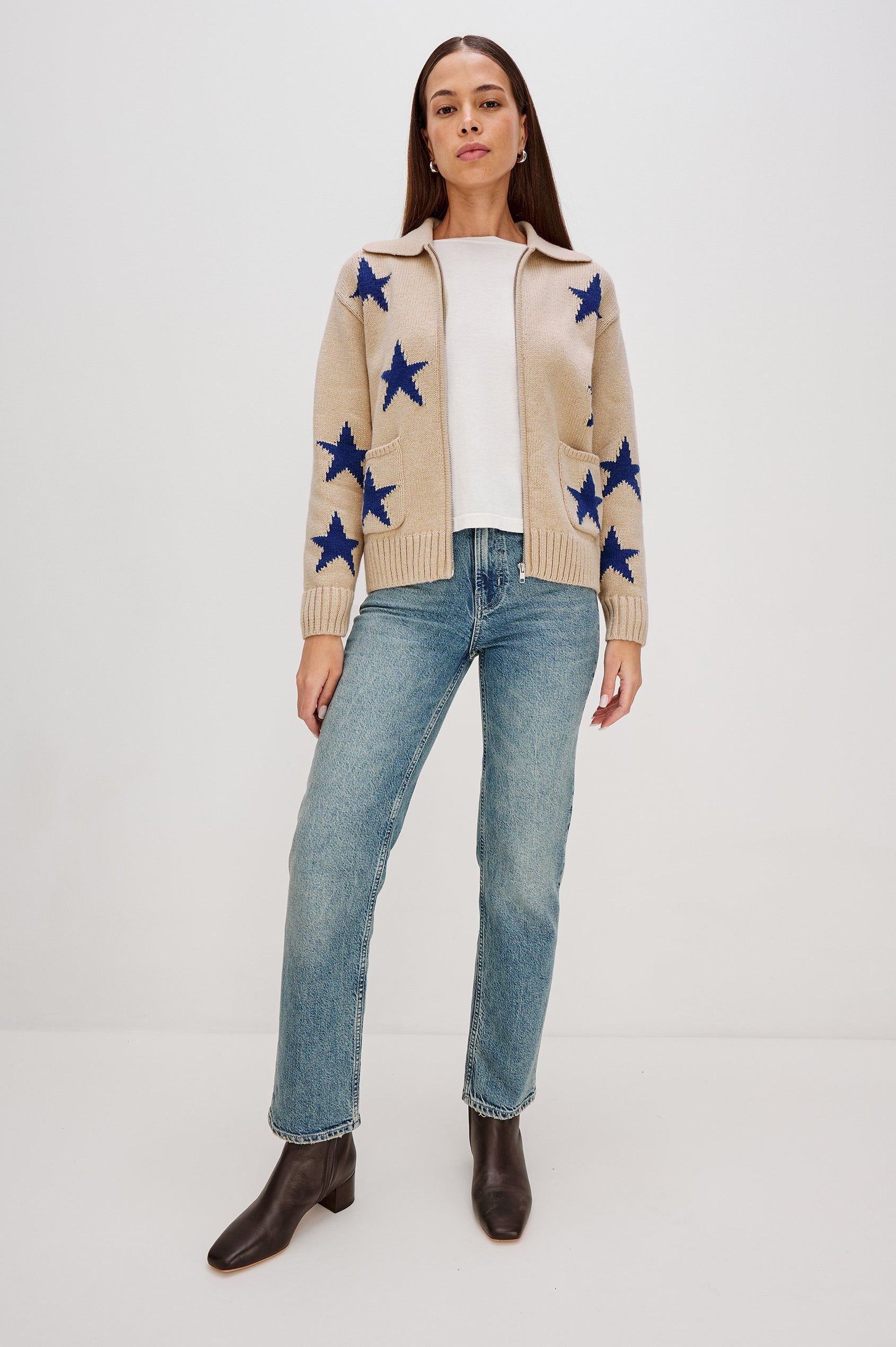 Rhea, a longline camel cardigan in a wool blend, featuring cobalt blue star motifs. The cardigan has a relaxed fit, zip-front closure, and front patch pockets. Full Body Front View Featured on Model, Unzipped