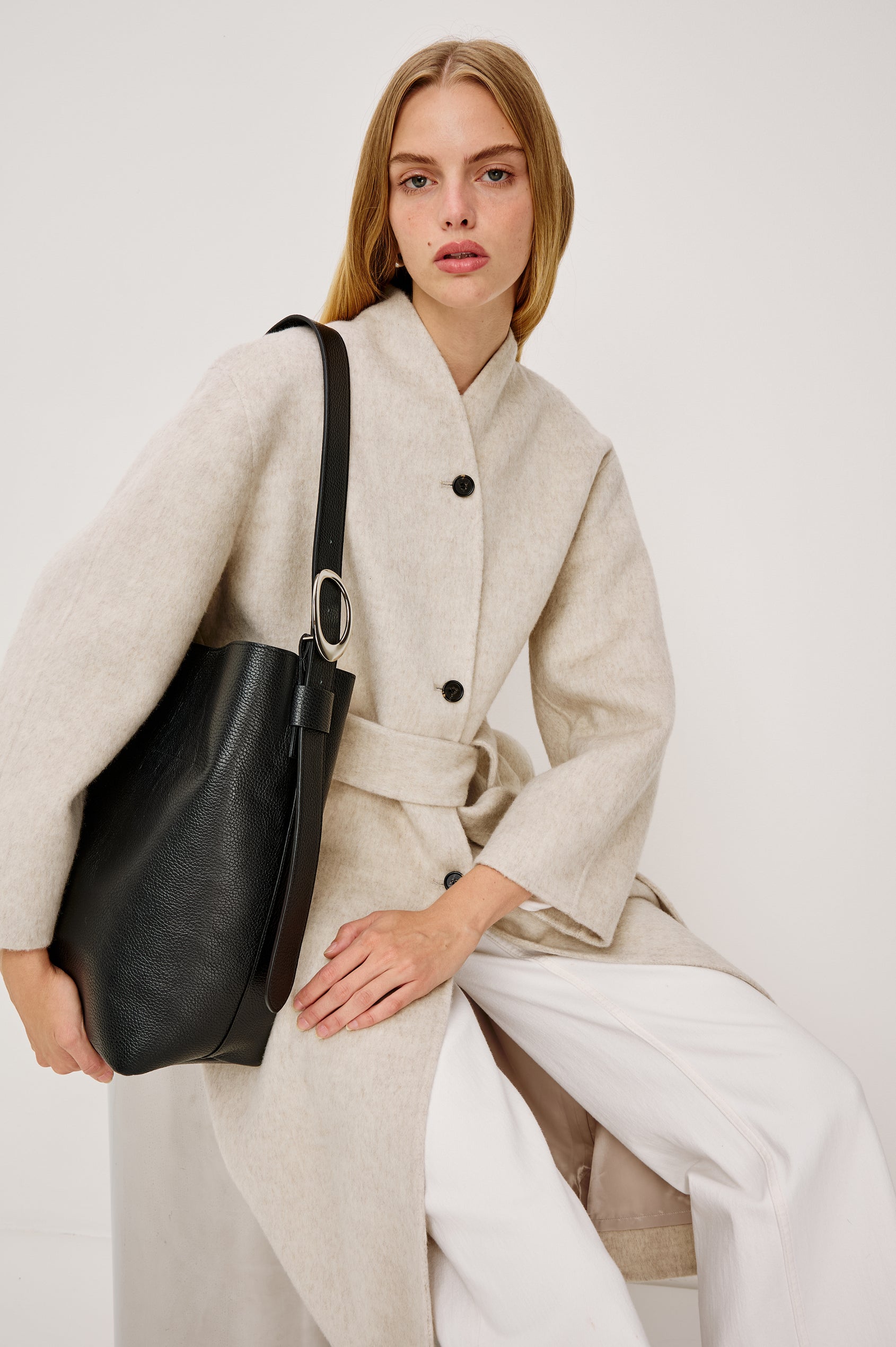 Roland Longline Coat in Wool Blend and Relaxed Fit With Slight Stand Collar, Front Buttons, Long Sleeves, Side Pockets, and an Adjustable Waist Belt in Oatmeal Colorway - Upper Body View Featured on Model Sitting