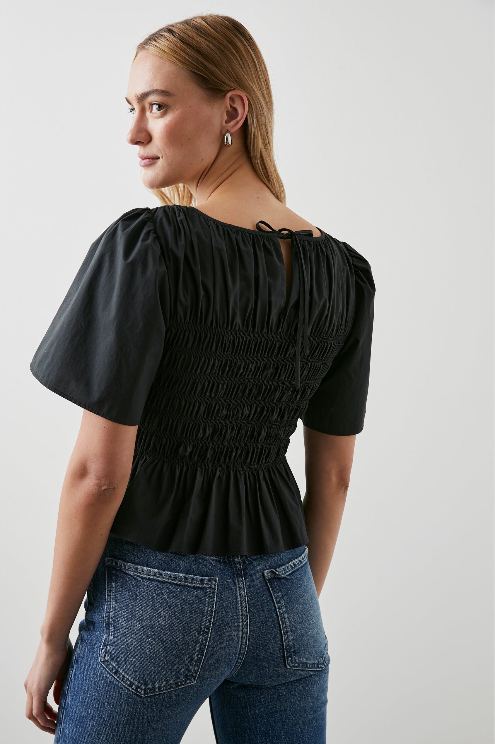 Rosie Short-Sleeve Top with a Smocked Center Bodice, Pleats at the Hem and Yoke and a Upper Back Tie Closure in Black Colorway - Back View Featured on Model