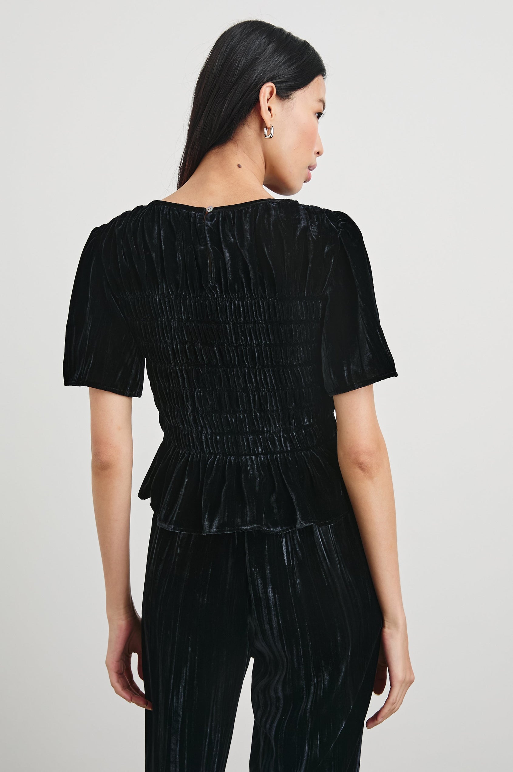 Rosie Short-Sleeve Top with a Smocked Center Bodice, Pleats at the Hem and Yoke and a Upper Back Button Closure in Black Velvet Colorway - Back View Featured on Model