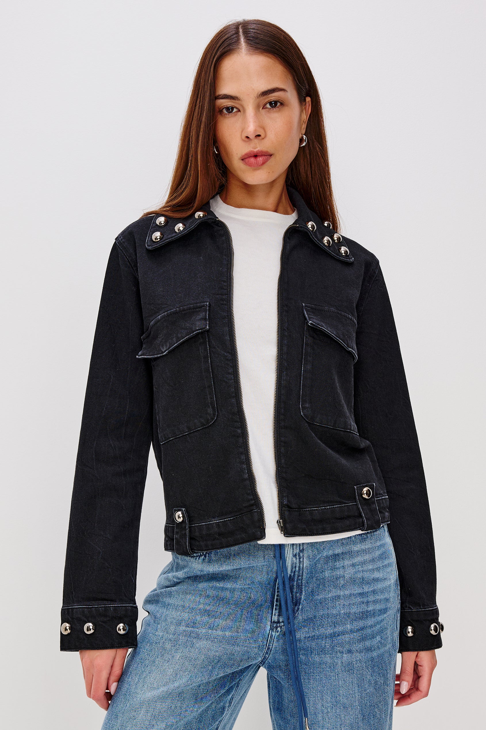 Roxbury is a boxy fit denim jacket featuring nail-back studs at the oversized collar, cuffs, and hem. It has oversized chest pockets with hidden snap closures, hem tabs, and a sleek front zipper closure in Black Stone Studded colorway. Front view featured on model.