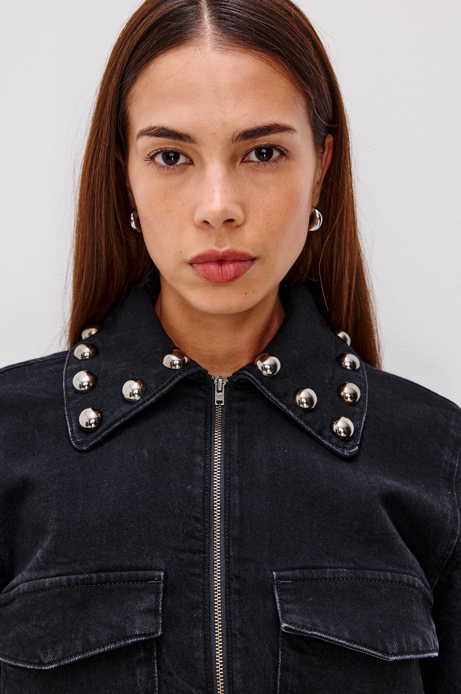 Roxbury Jacket in Denim With Boxy Fit, Nail-Back Studs at Oversized Collar, Long Sleeve Cuffs, and Hem, Oversized Chest Pockets With Hidden Snap Closures, Hem Tabs, and Front Zip in Black Stone Studded Colorway - Upper Body Front Close Up View Featured on Model