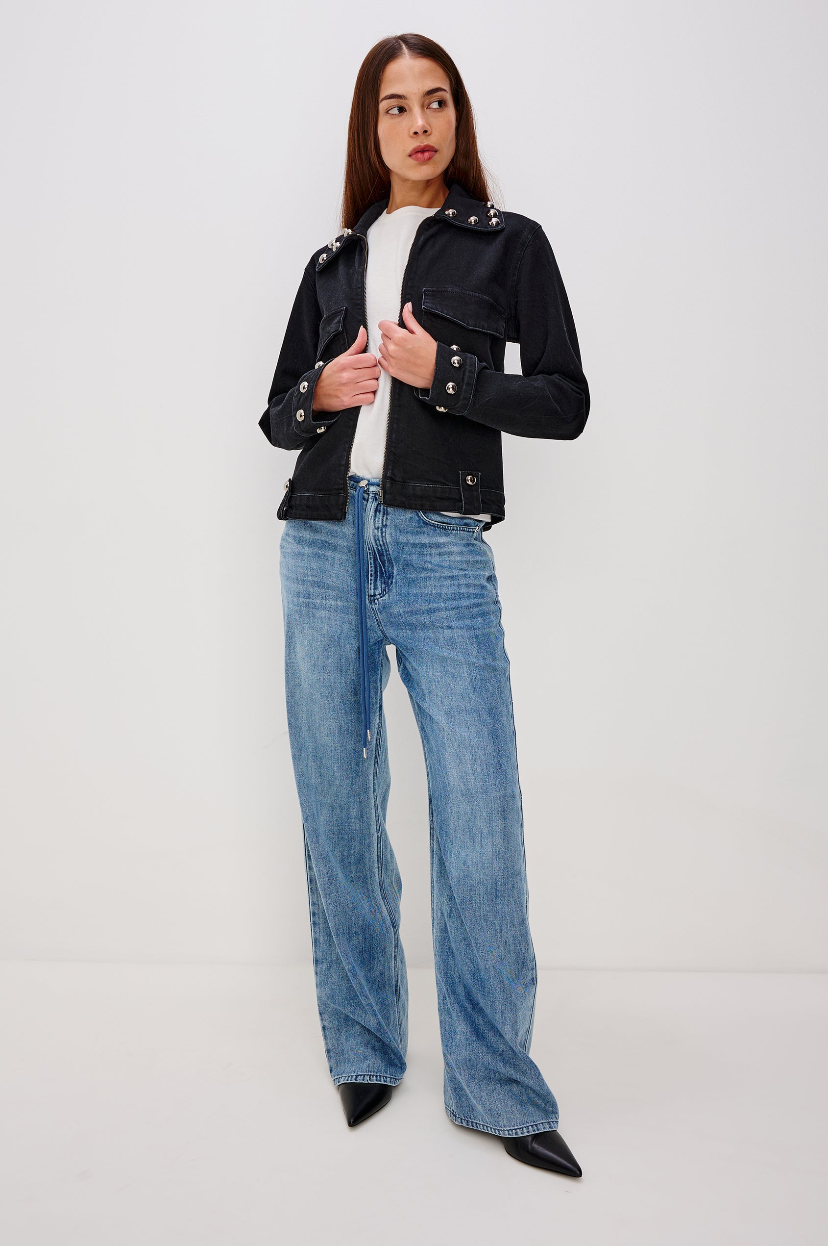 Roxbury is a boxy fit denim jacket featuring nail-back studs at the oversized collar, cuffs, and hem. It has oversized chest pockets with hidden snap closures, hem tabs, and a sleek front zipper closure in Black Stone Studded colorway. Front full body view featured on model.