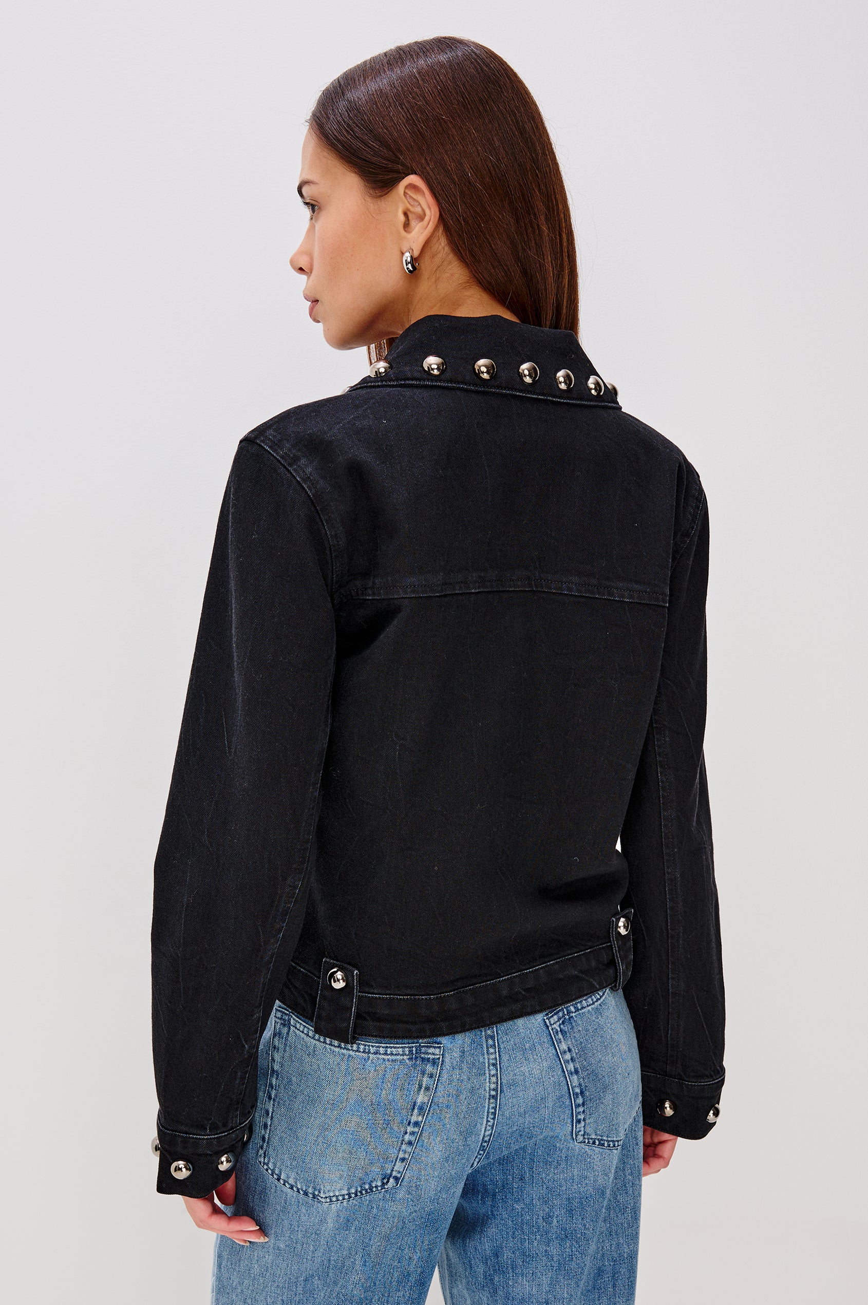 Roxbury is a boxy fit denim jacket featuring nail-back studs at the oversized collar, cuffs, and hem. It has oversized chest pockets with hidden snap closures, hem tabs, and a sleek front zipper closure in Black Stone Studded colorway. Back view featured on model.