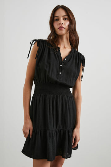 Samina Above-Knee Sleeveless Dress with drawstrings at the Sleeve Openings, Buttons along the Bodice, A Cinched Waist and a Tiered Skirt in Black Colorway - Front View Featured on Model 