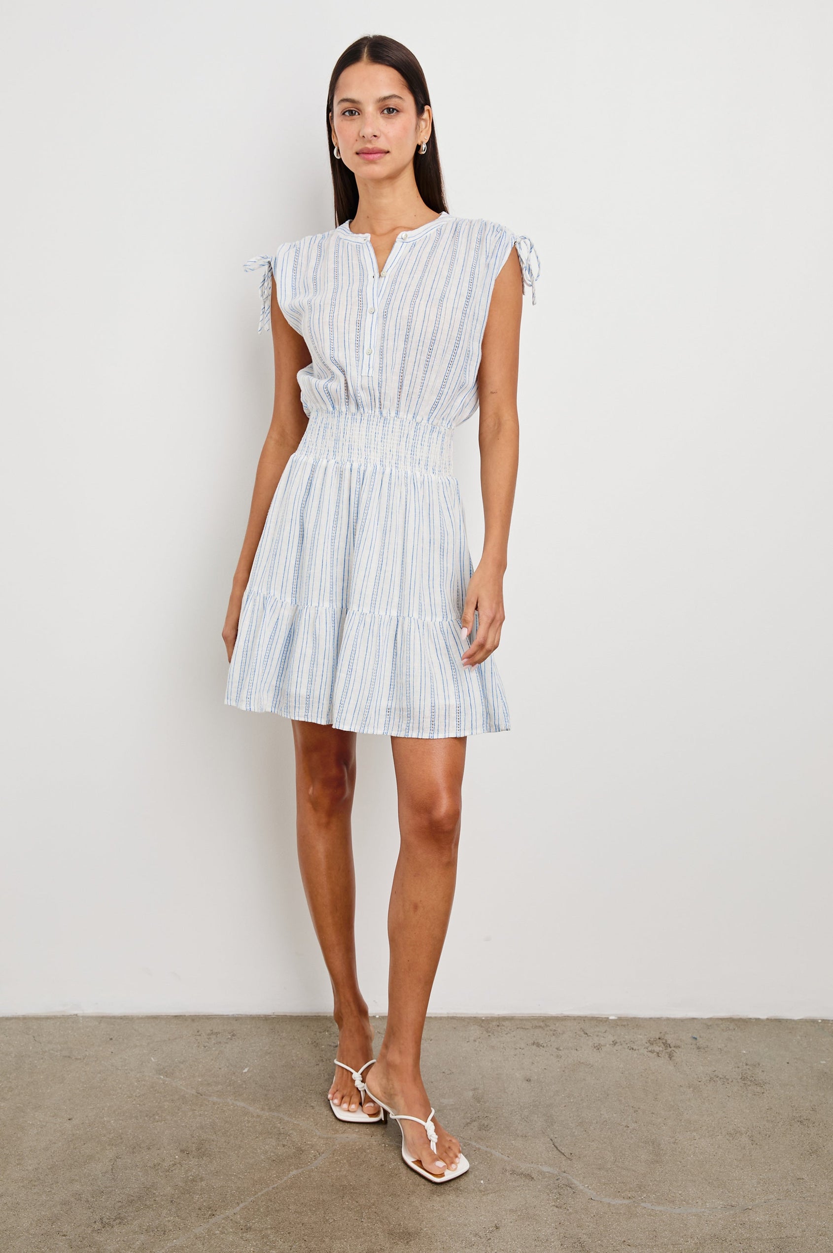 Samina Above-Knee Sleeveless Dress with drawstrings at the Sleeve Openings, Buttons along the Bodice, A Cinched Waist and a Tiered Skirt in Sonoma Stripe Colorway, Thin Vertical Blue Stripes on White - Front Full Body View Featured on Model 