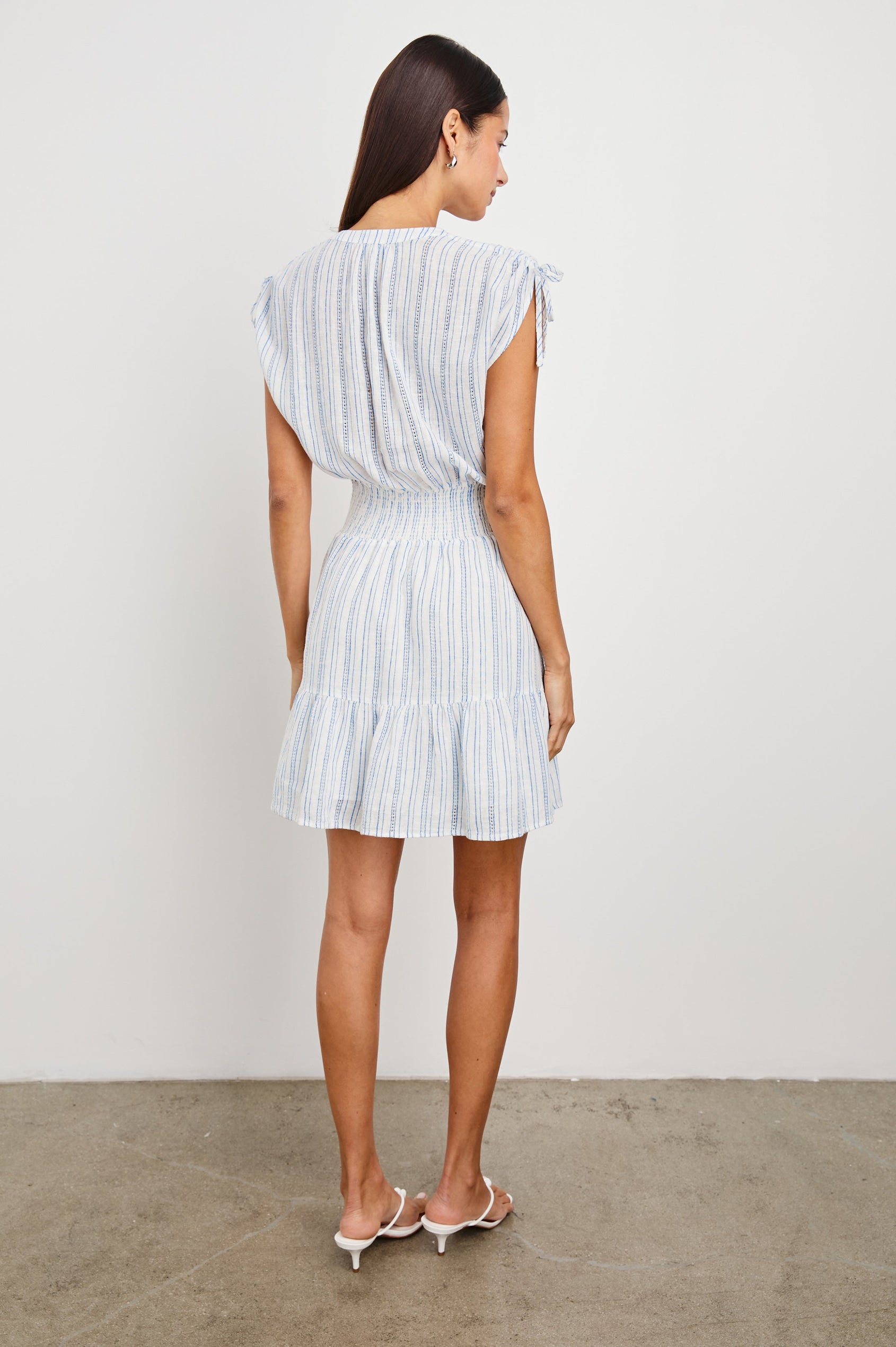Samina Above-Knee Sleeveless Dress with drawstrings at the Sleeve Openings, Buttons along the Bodice, A Cinched Waist and a Tiered Skirt in Sonoma Stripe Colorway, Thin Vertical Blue Stripes on White - Back Full Body View Featured on Model 