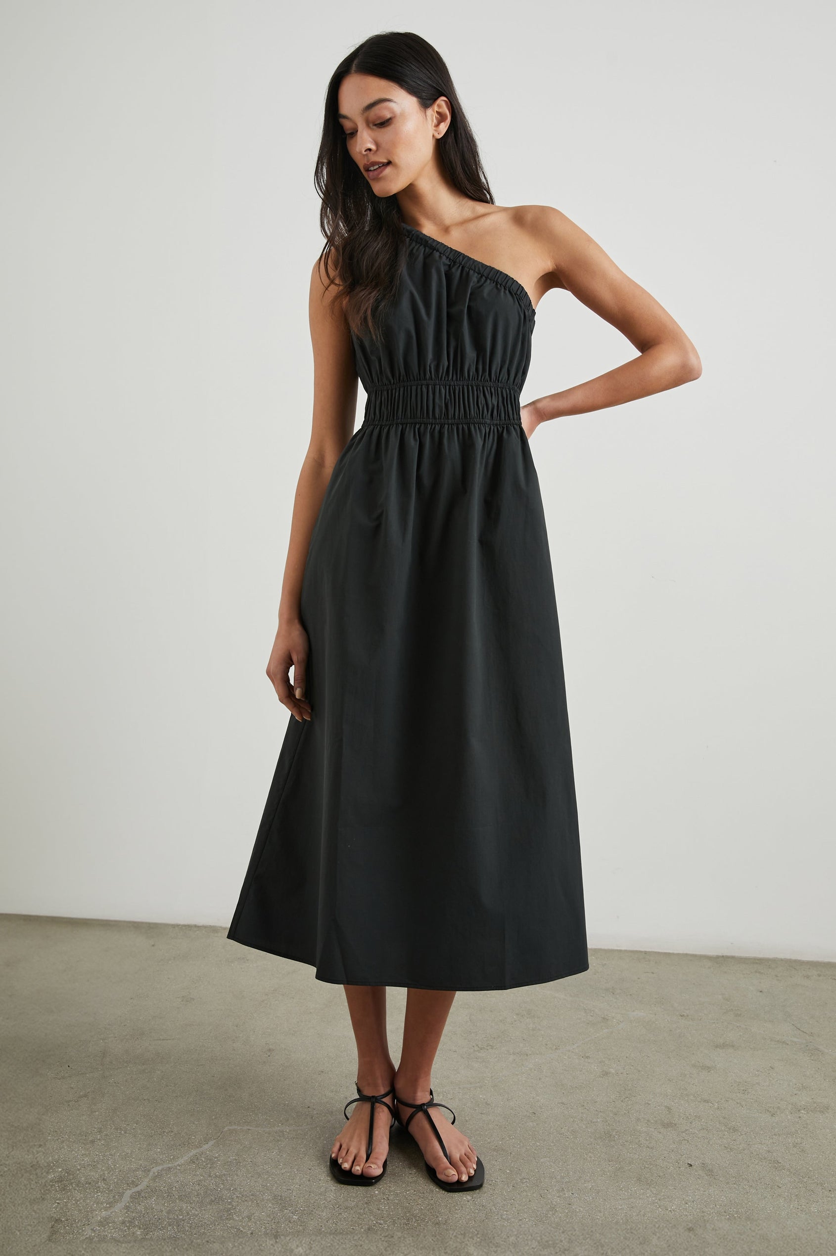 Selani Midi Dress With an Asymmetrical Neckline, Mid-Thigh Slide Slit and Cinched Waistband in Black Colorway - Front Full Body View Featured on Model