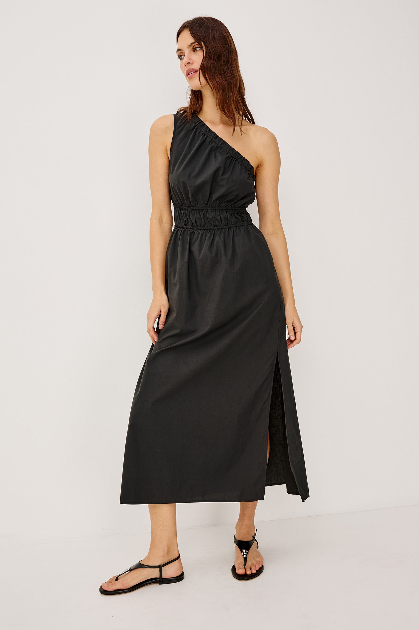 Selani Midi Dress With an Asymmetrical Neckline, Mid-Thigh Slide Slit and Cinched Waistband in Black Colorway - Front Full Body View Featured on Model