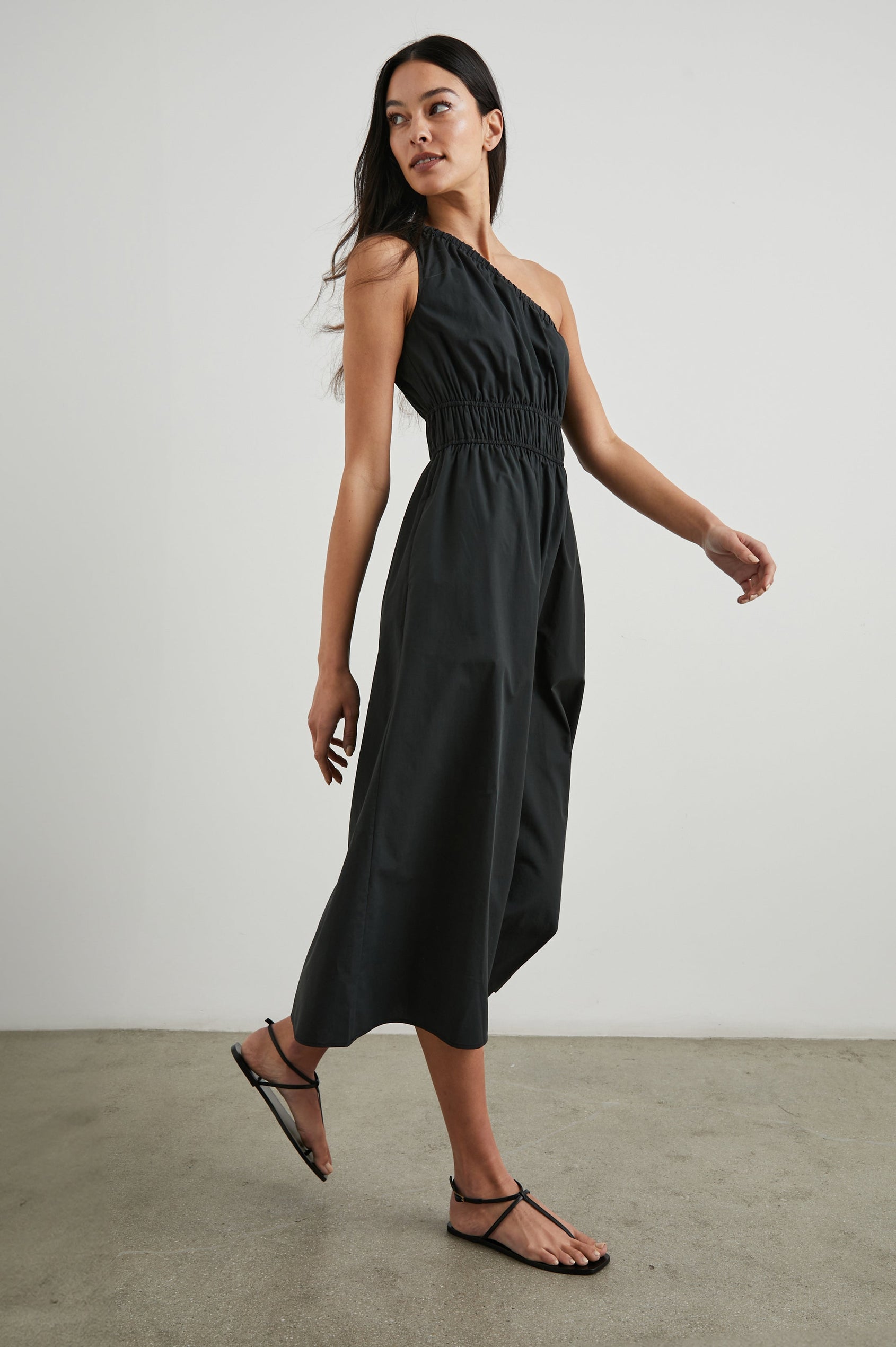 Selani Midi Dress With an Asymmetrical Neckline, Mid-Thigh Slide Slit and Cinched Waistband in Black Colorway - Side Full Body View Featured on Model