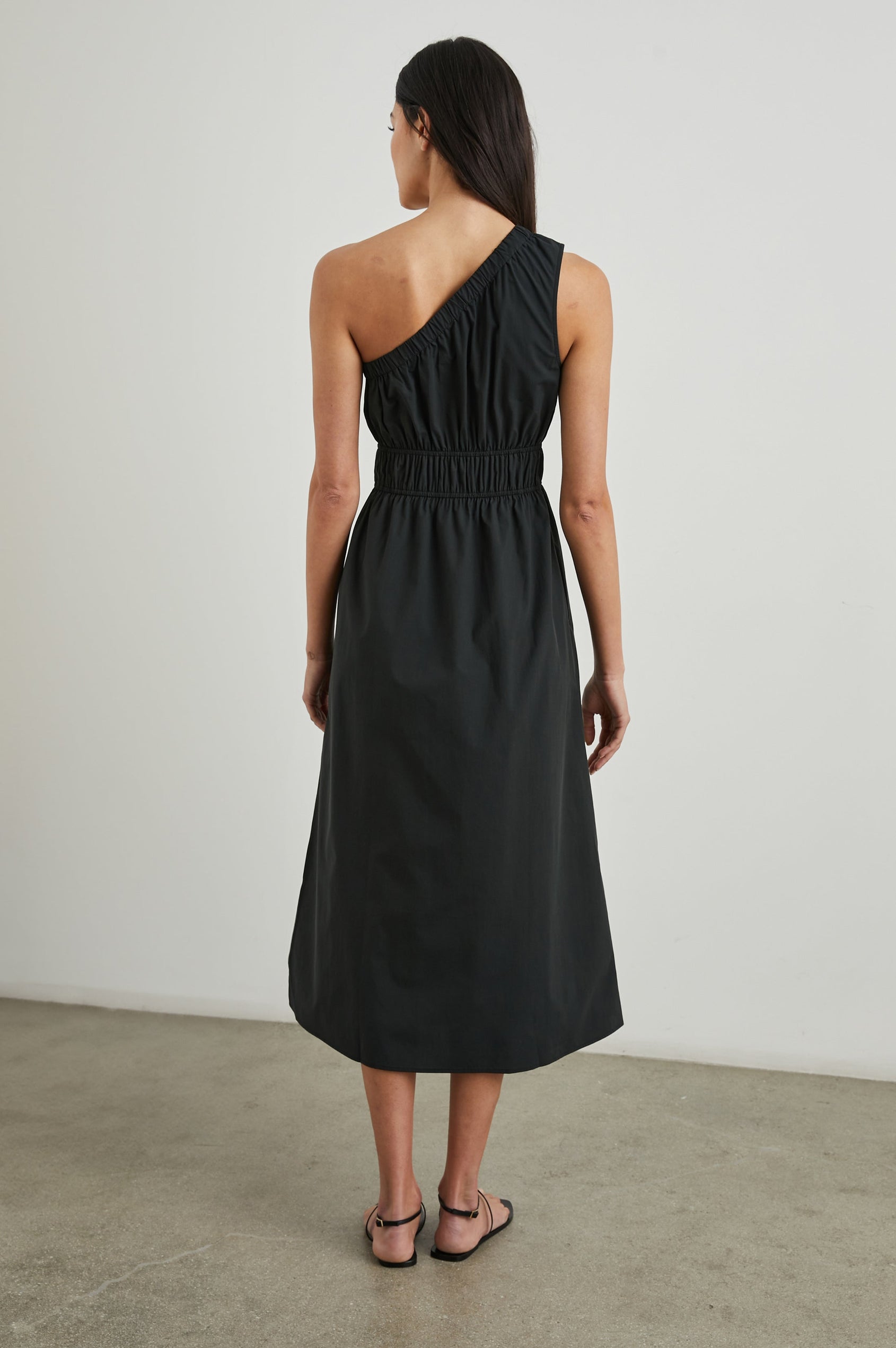 Selani Midi Dress With an Asymmetrical Neckline, Mid-Thigh Slide Slit and Cinched Waistband in Black Colorway - Back Full Body View Featured on Model