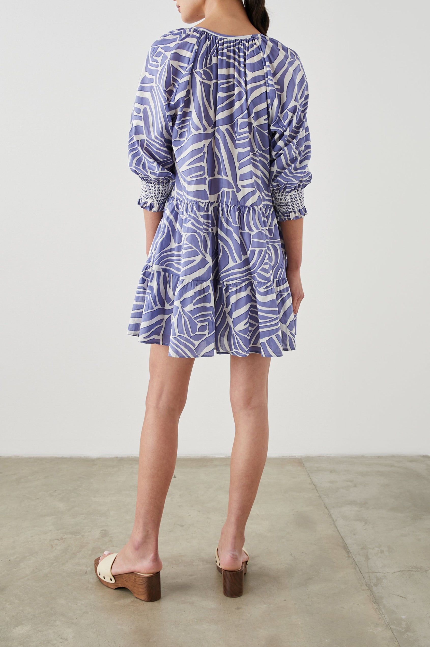 Sia Mini Dress with Three-Quarter Sleeves, Shirred Cuffs, a Tiered Skirt, and a V Neck with a Tie and Light Ruching in Island Waves Colorway, a Purple and White Print - Back Full Body View Featured on Model 