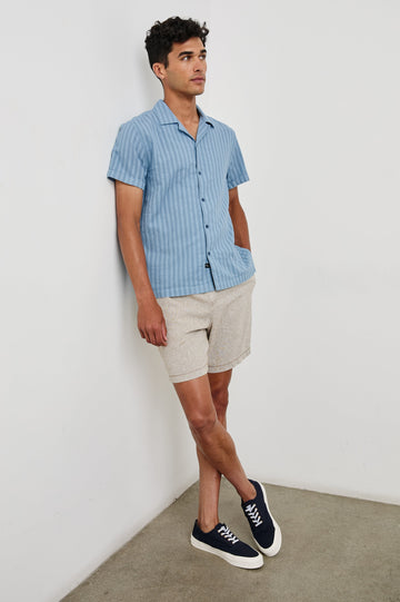 Sinclair short sleeve button up shirt with camp collar in Faded Blue with stripes - full body front view on model, leaning on wall