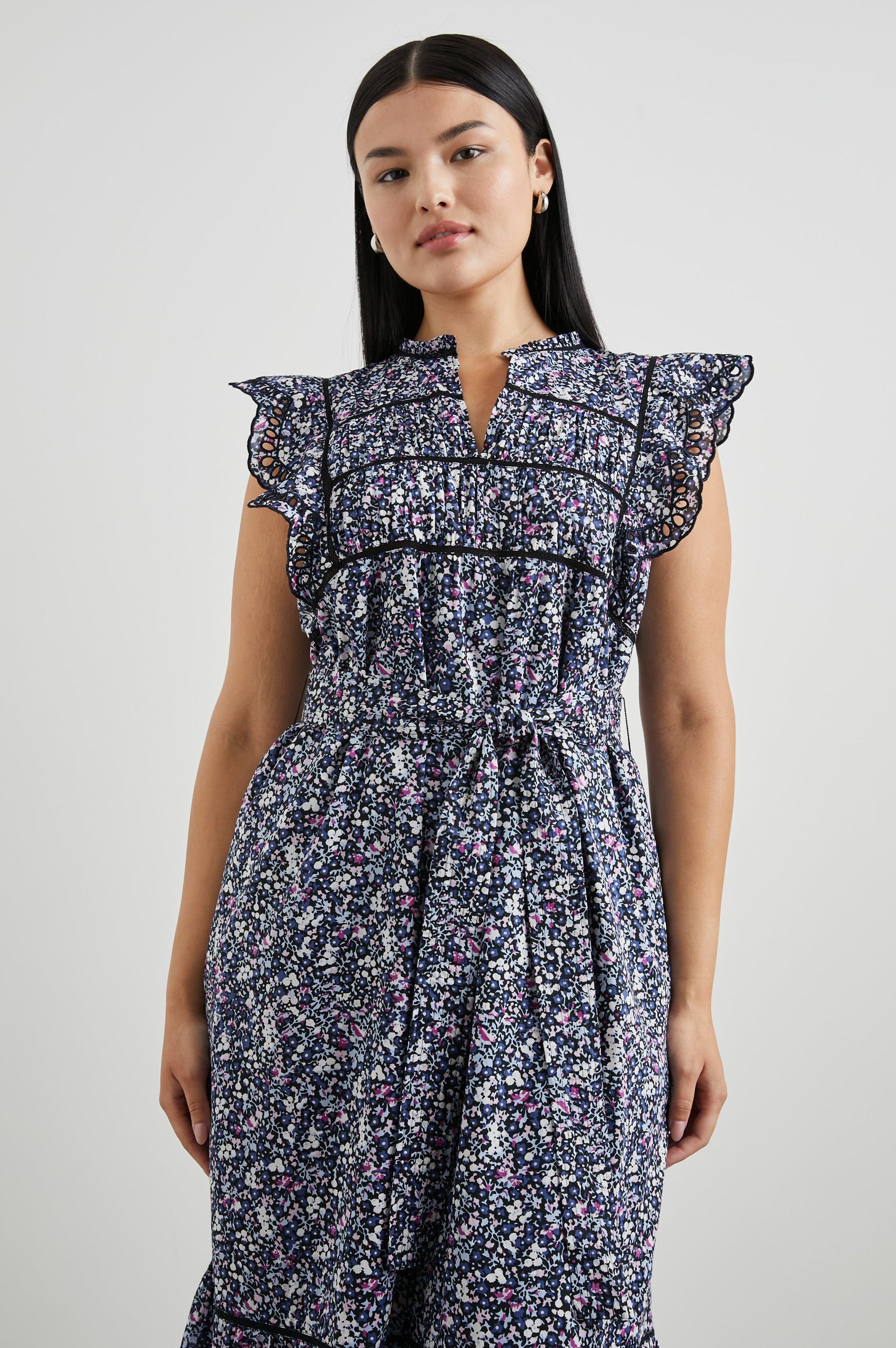 Sofie Short Sleeve Midi Dress with Flutter Sleeves, a V Neck, Panels with Shirring on the Bodice, a Fabric Waist Tie, a Tiered Skirt, and Cutouts on the Sleeves and Along the Hem in Midnight Hyacinth Colorway, Small Flowers in Blues, Pinks and White on Midnight - Front View Featured on Model 