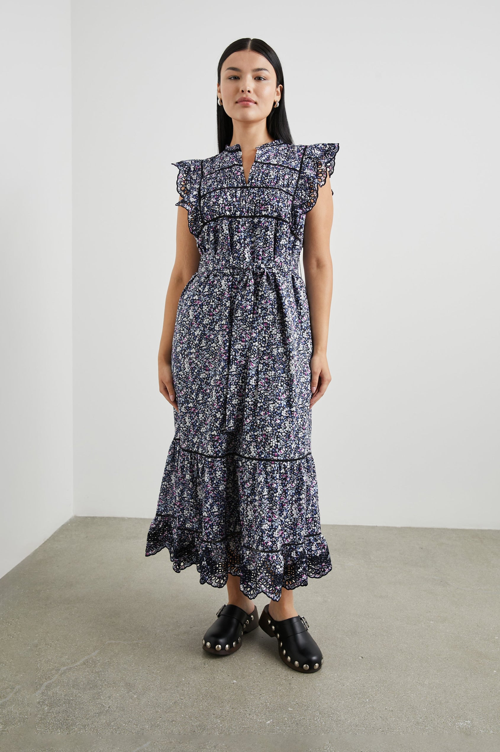 Sofie Short Sleeve Midi Dress with Flutter Sleeves, a V Neck, Panels with Shirring on the Bodice, a Fabric Waist Tie, a Tiered Skirt, and Cutouts on the Sleeves and Along the Hem in Midnight Hyacinth Colorway, Small Flowers in Blues, Pinks and White on Midnight - Front Full Body View Featured on Model 