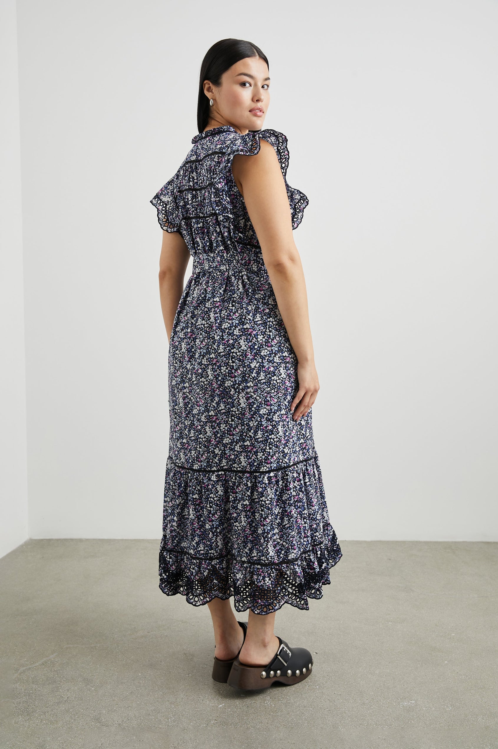 Sofie Short Sleeve Midi Dress with Flutter Sleeves, a V Neck, Panels with Shirring on the Bodice, a Fabric Waist Tie, a Tiered Skirt, and Cutouts on the Sleeves and Along the Hem in Midnight Hyacinth Colorway, Small Flowers in Blues, Pinks and White on Midnight - Back Full Body View Featured on Model 