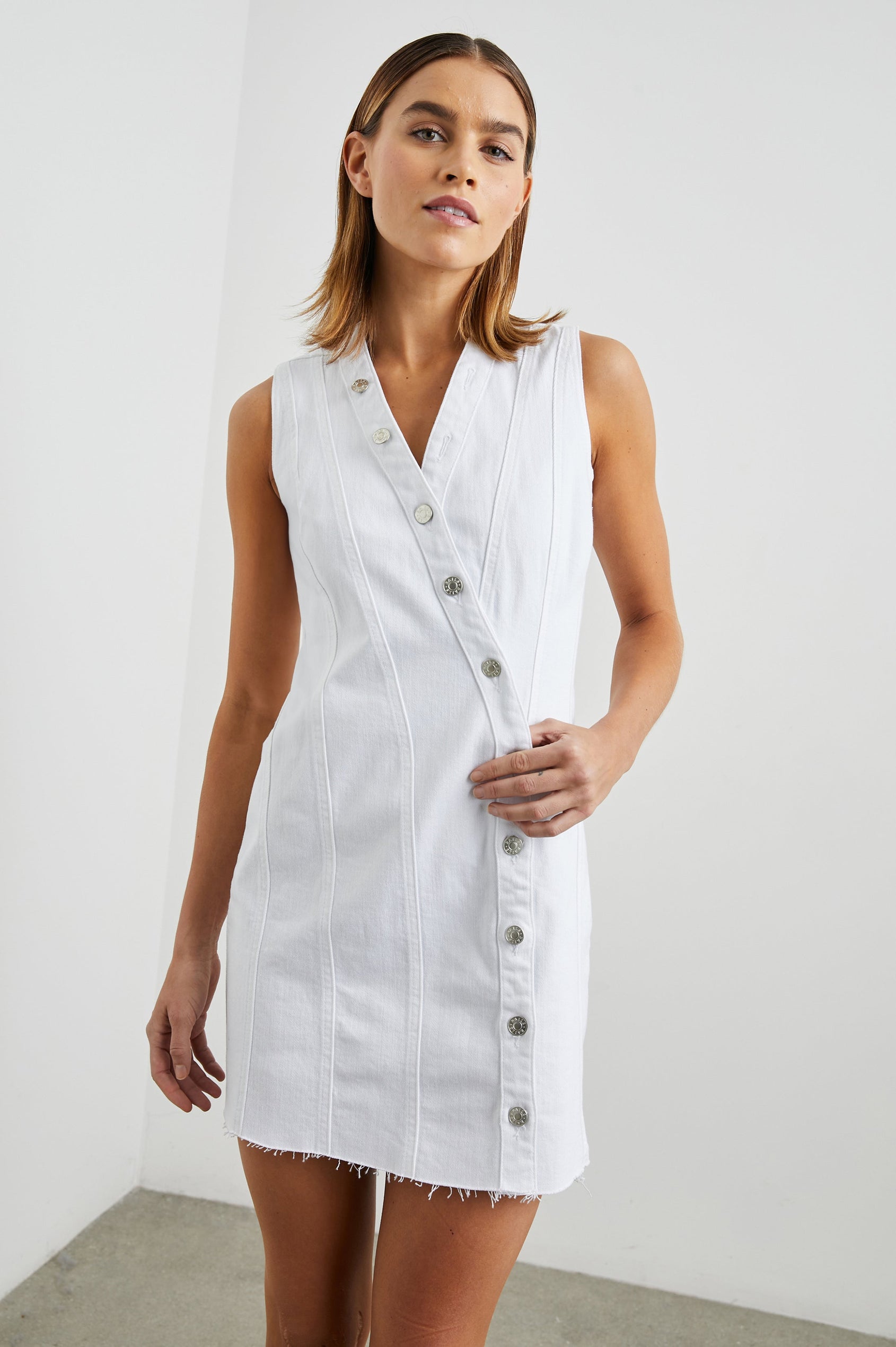 Strand Sleeveless Denim Mini Dress with a V Neck, a Raw Hem, Wide Shoulder Straps, a Row of Buttons that Extend Diagonally Across the Bodice and then Vertically Down the Skirt, Vertical Seams in Salt White Colorway - Front View Featured on Model  