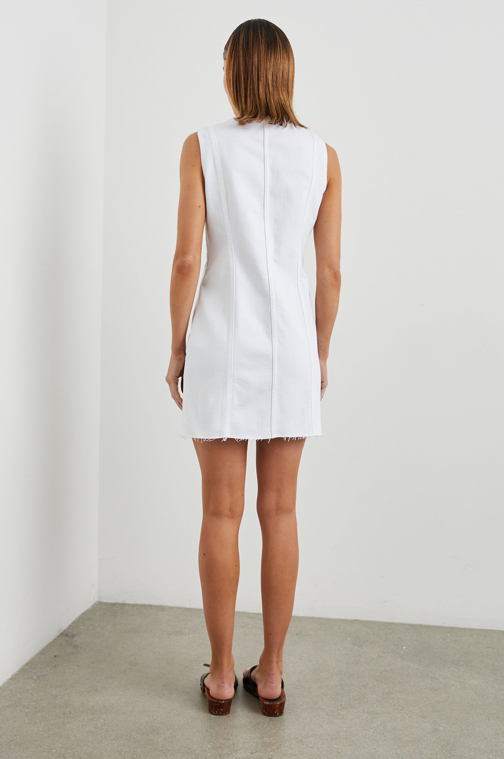 Strand Sleeveless Denim Mini Dress with a V Neck, a Raw Hem, Wide Shoulder Straps, a Row of Buttons that Extend Diagonally Across the Bodice and then Vertically Down the Skirt, Vertical Seams in Salt White Colorway - Back Full Body View Featured on Model  