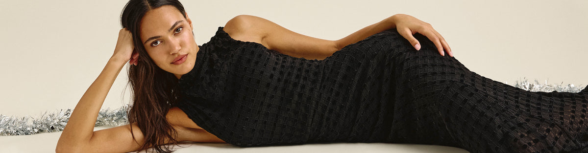 Woman reclining on her side in a sleeveless black textured maxi dress, propping her head on one hand, with silver tinsel garland behind her against a cream backdrop.