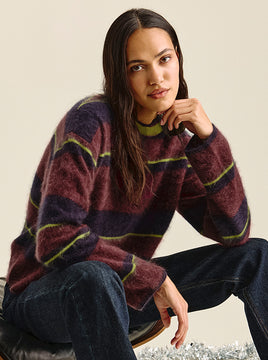 Woman with long dark hair seated and resting her chin on her hand, wearing a fuzzy striped sweater in burgundy and deep purple with thin lime-green accent lines, paired with dark denim jeans.