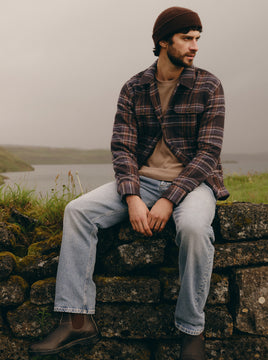 A man wearing a brown knit beanie, a brown and black plaid flannel shirt over a tan t-shirt, light blue jeans, and brown boots sits on a mossy stone wall outdoors with a lake and grassy hills in the background.