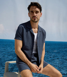 Man sitting on a chair by the ocean wearing a navy blue outfit.