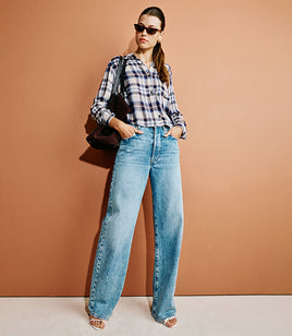 A woman stands against a warm tan wall wearing a black-and-white plaid button-up shirt, light blue wide-leg jeans, and clear-strap sandals, with a black shoulder bag.