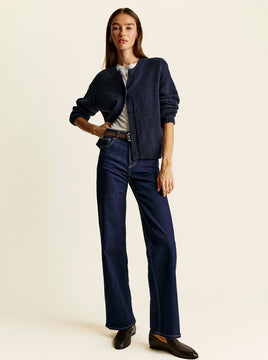 Woman with long brown hair stands against a cream background, wearing a navy knit button-up cardigan over a white tee, dark rinse wide-leg jeans with contrast stitching, a brown belt, and black leather loafers, one hand on her hip.