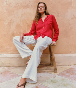 Woman in a red shirt and white pants sitting on a wooden stool against a textured wall.