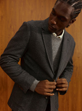 A man wearing a dark gray wool blazer over a gray knit sweater and white collared shirt, standing against a warm wood-paneled background.