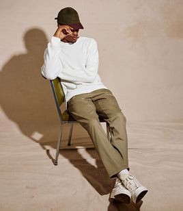 A model sits on a green and metal chair against a textured beige wall, wearing a white long-sleeve knit sweater, olive green trousers, white sneakers, and a dark cap.