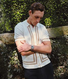Man wearing a white shirt with brown accents and a watch, standing against a natural background.