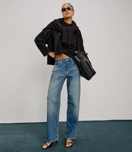 Person wearing a black outfit with blue jeans and sunglasses, holding a black bag against a plain background.