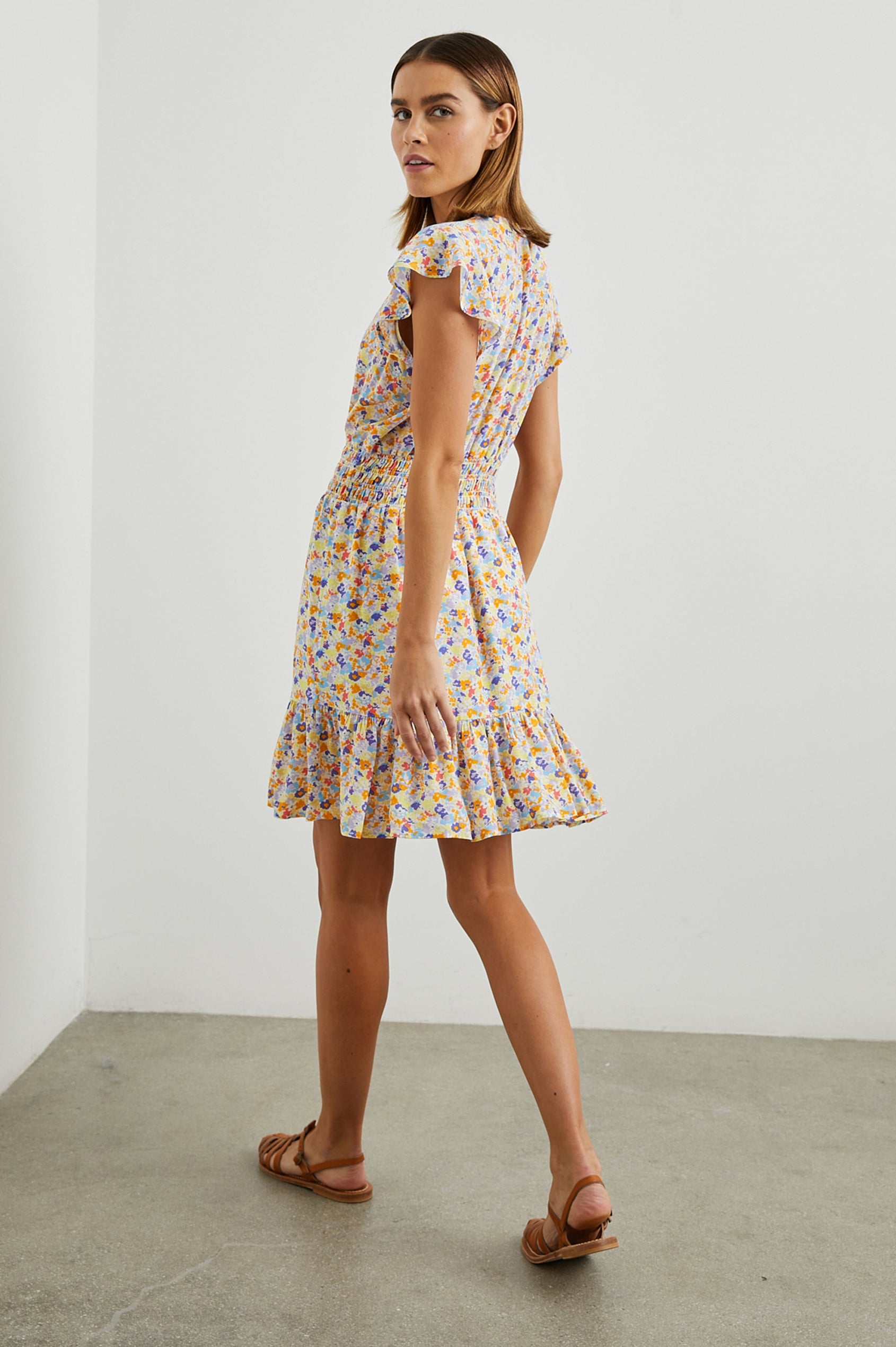 Tara Short Sleeve, Above the Knee Dress with a Smocked Waist, V Neck, Flutter Cap Sleeves, and Tiered Skirt with a Ruffled Hem in Papaya Flowers Colorway, in Orange, Purple, Yellow and Blue - Side Full Body View Featured on Model 