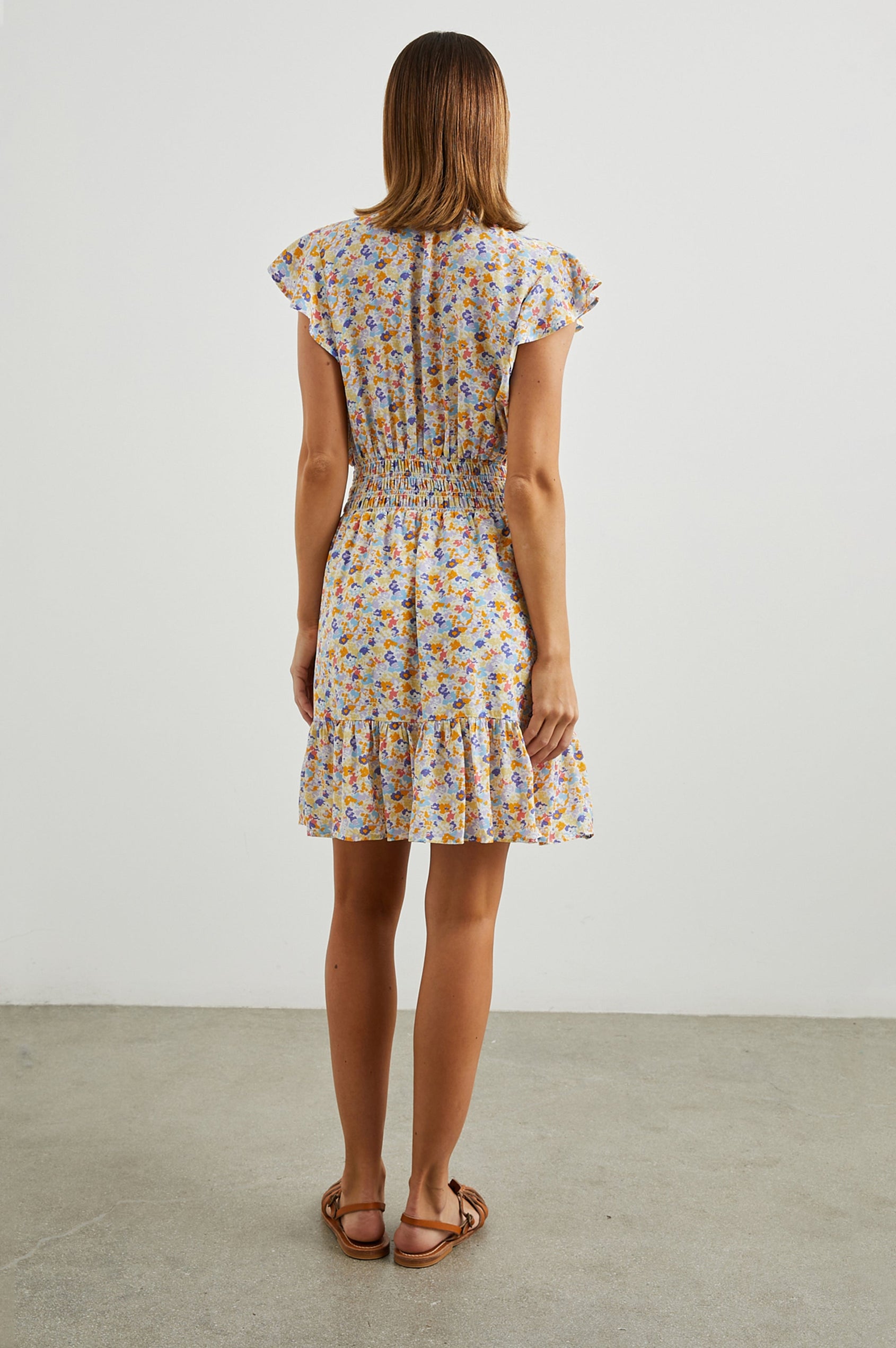 Tara Short Sleeve, Above the Knee Dress with a Smocked Waist, V Neck, Flutter Cap Sleeves, and Tiered Skirt with a Ruffled Hem in Papaya Flowers Colorway, in Orange, Purple, Yellow and Blue - Back Full Body View Featured on Model 