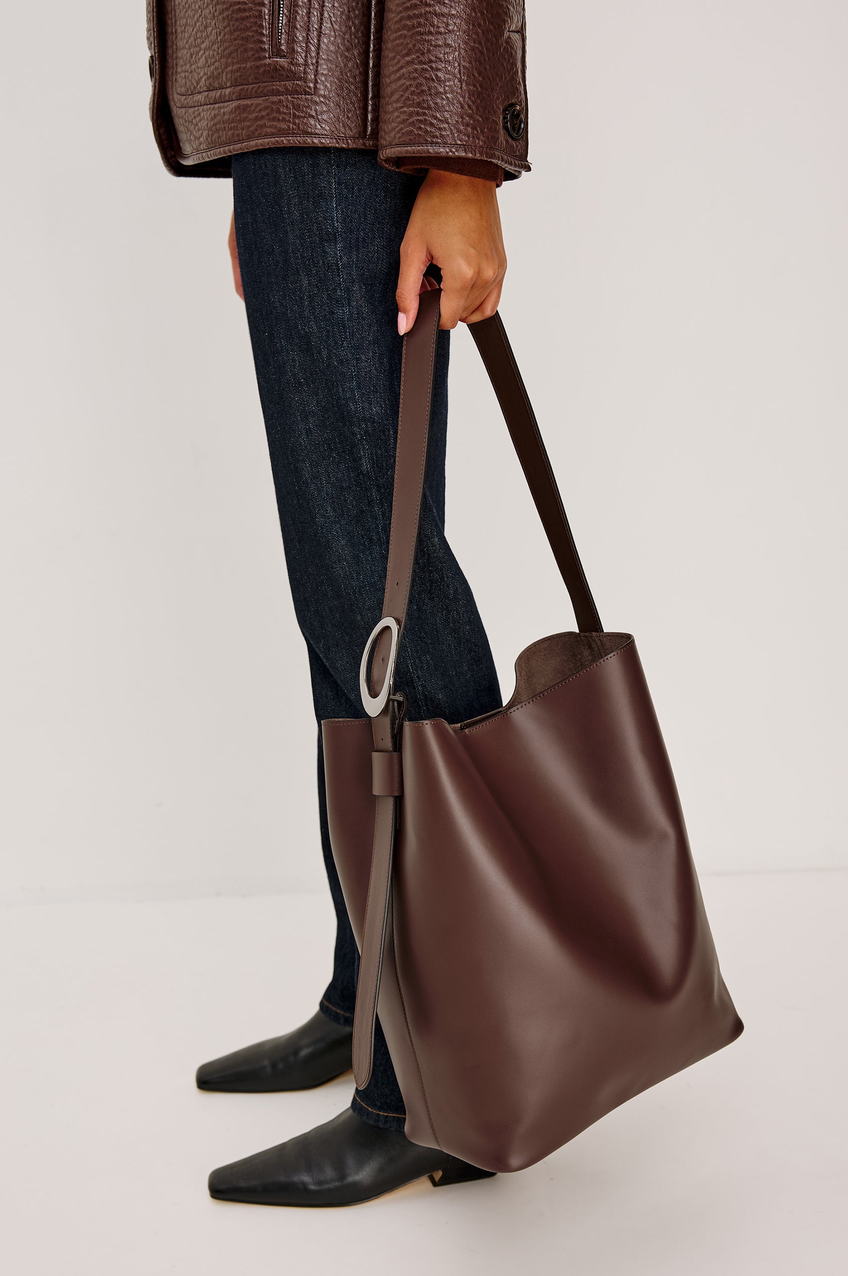 The Torino is a soft leather bucket bag, designed with an adjustable strap and polished silver oval buckle, in espresso colorway. Side view featured on model.