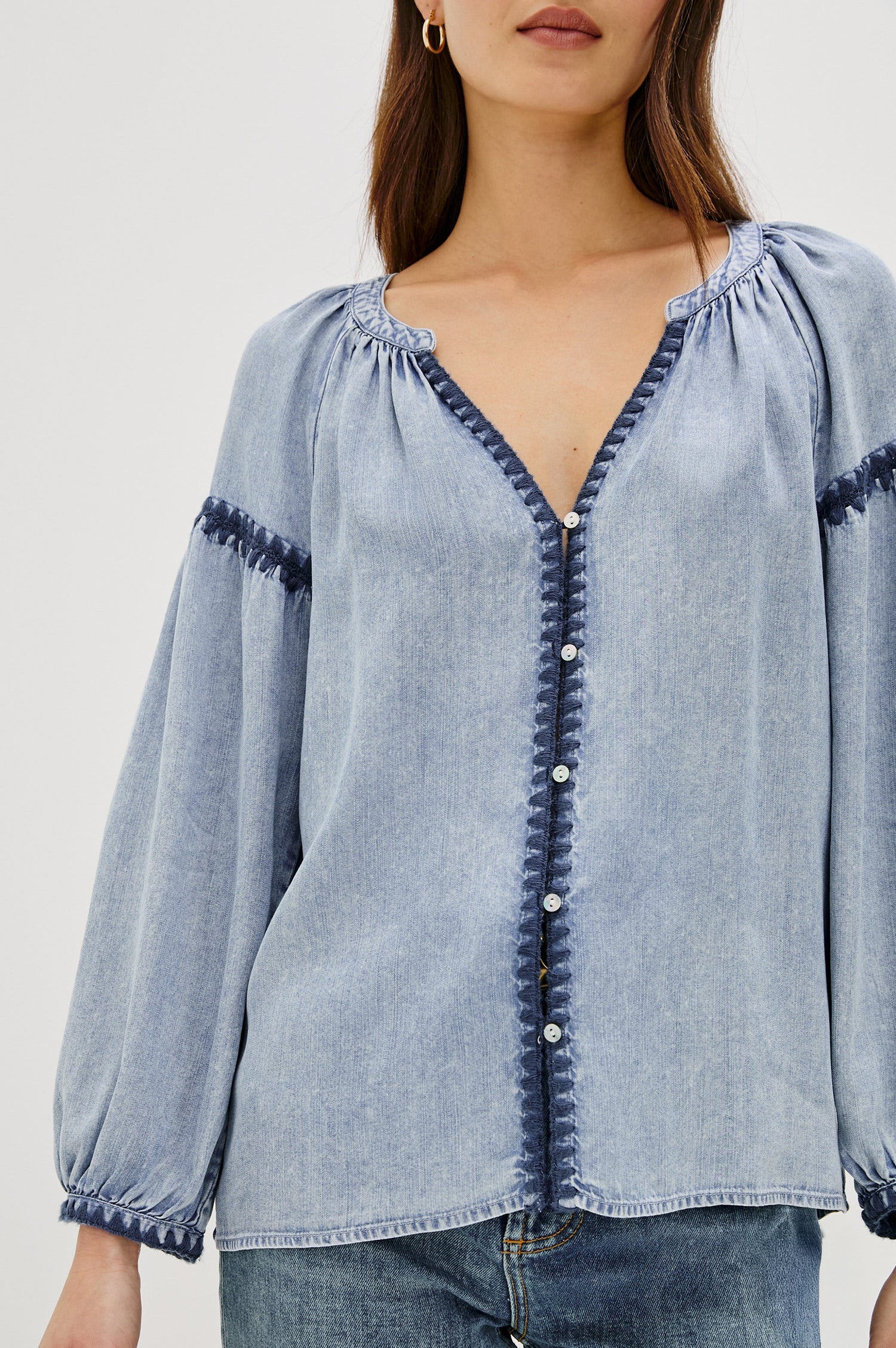 Toshi loose-fitting long sleeve top featuring a v-neckline, front button closure in Medium Cloud Wash Colorway. Contrast blanket stitching at the front, below shoulder sleeve and at the cuff - close up of details on model, untucked