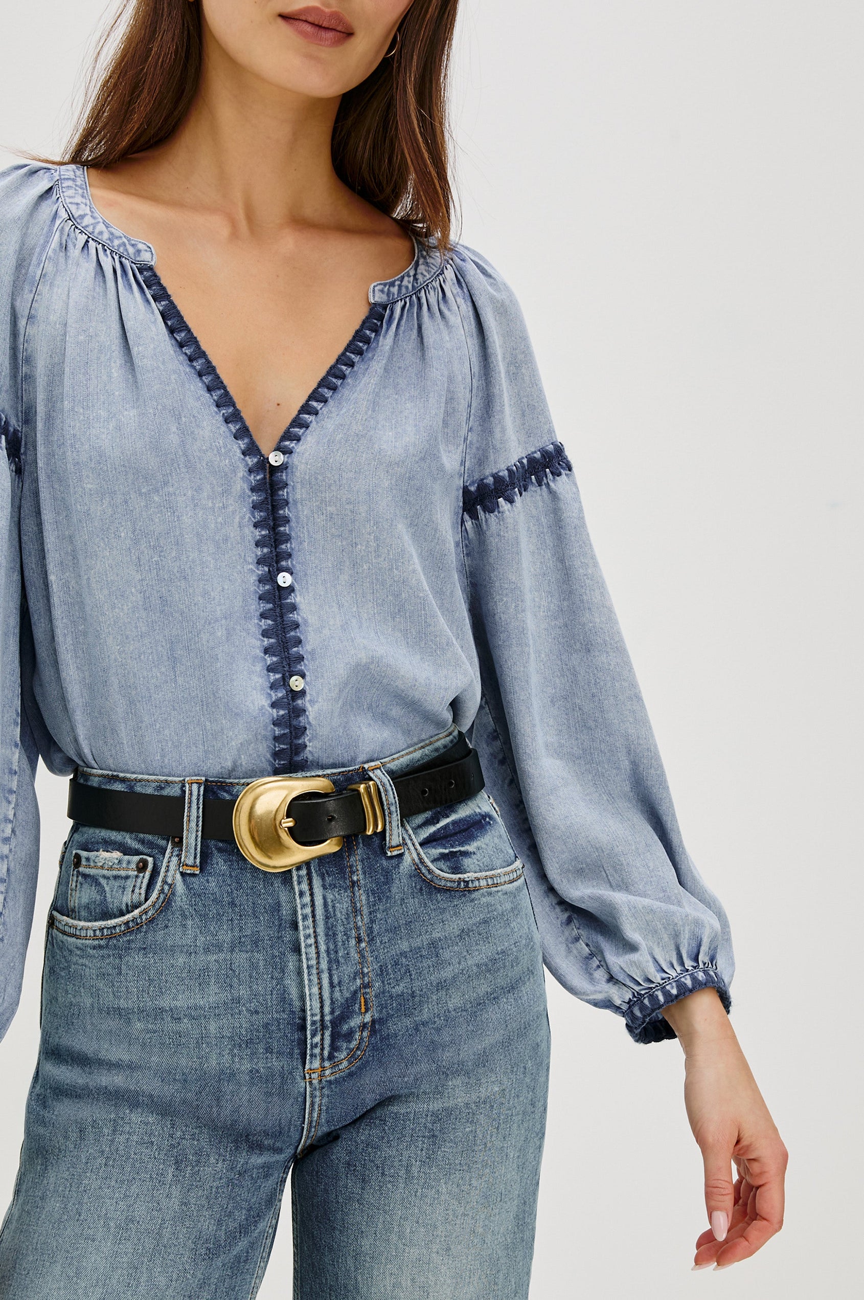 Toshi loose-fitting long sleeve top featuring a v-neckline, front button closure in Medium Cloud Wash Colorway. Contrast blanket stitching at the front, below shoulder sleeve and at the cuff - close up of details on model, shirt tucked in with belt