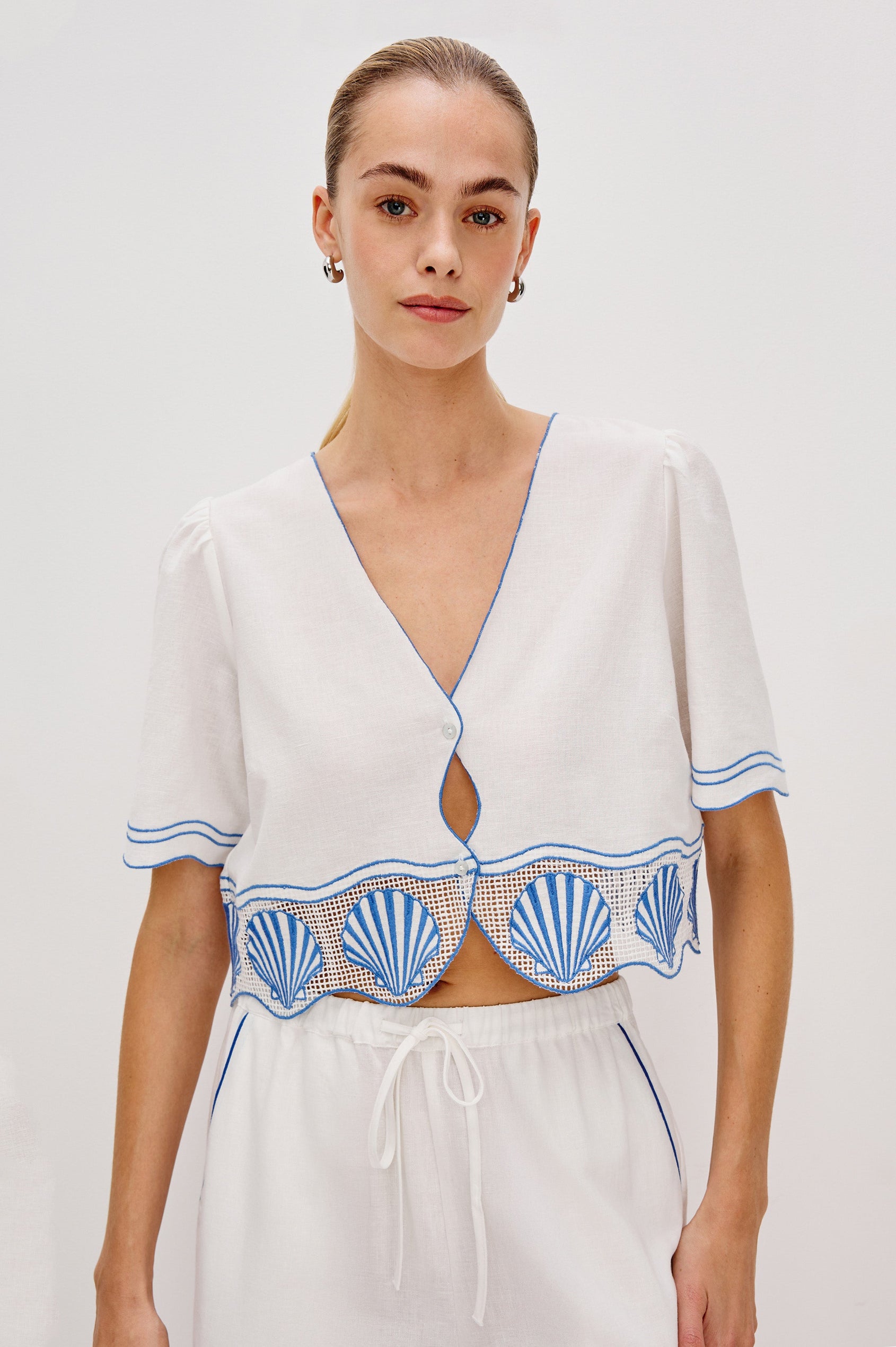 Venice Cropped V-Neck Top with Short Sleeves (slightly puffed), 2-Button Front, Keyhole at Center Front, Contrast Blue Sea Shell Embroidery and Scalloped Hem in White Colorway. Front View Shown on Model.