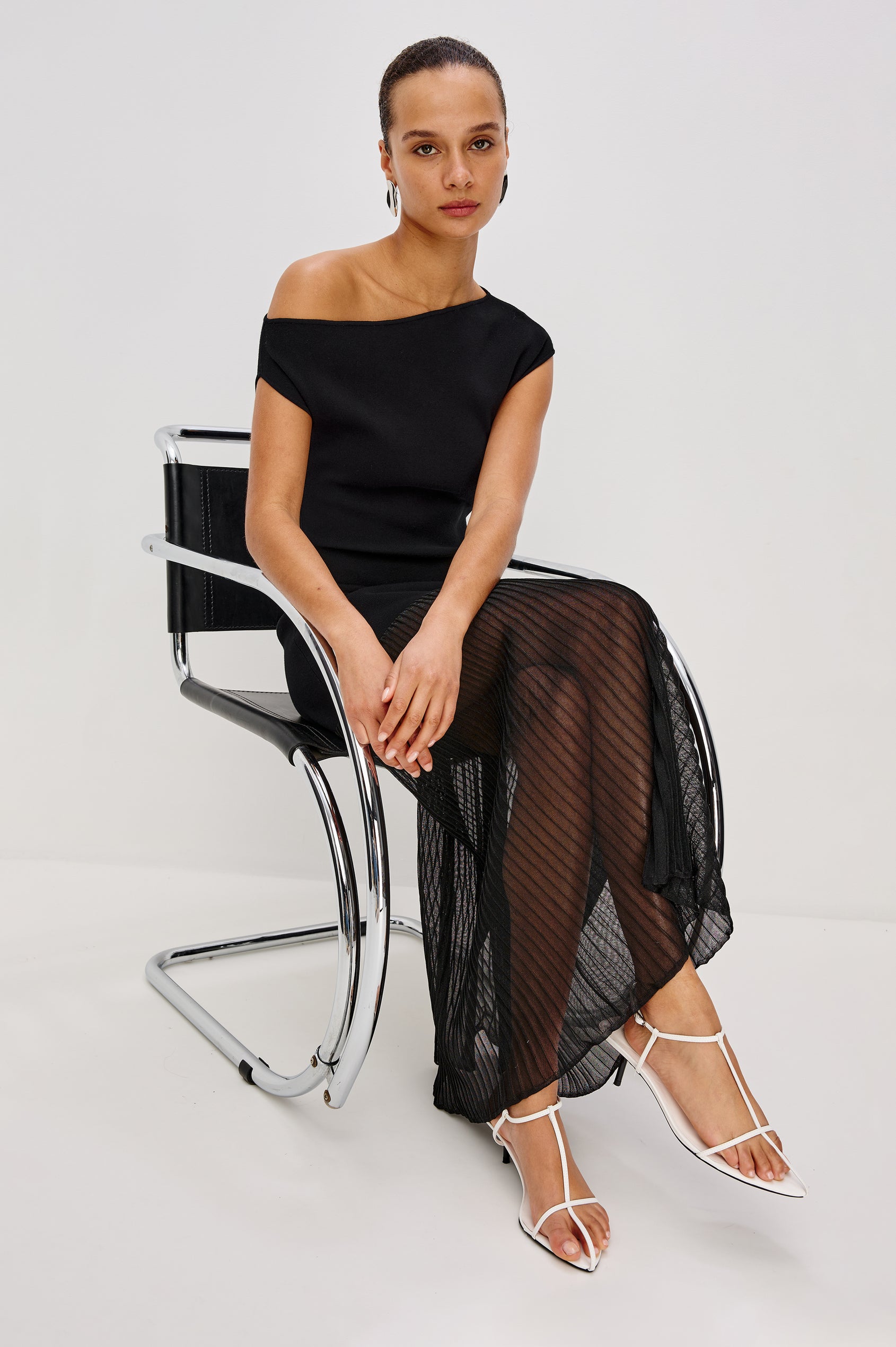 A fitted black midi dress with an off-shoulder neckline. The compact rib knit hugs the body through the hips, then transitions into a sheer pleated organza panel that finishes at a flowing midi hem. Full Body Front View featured on Model sitting in black leather and metal chair.