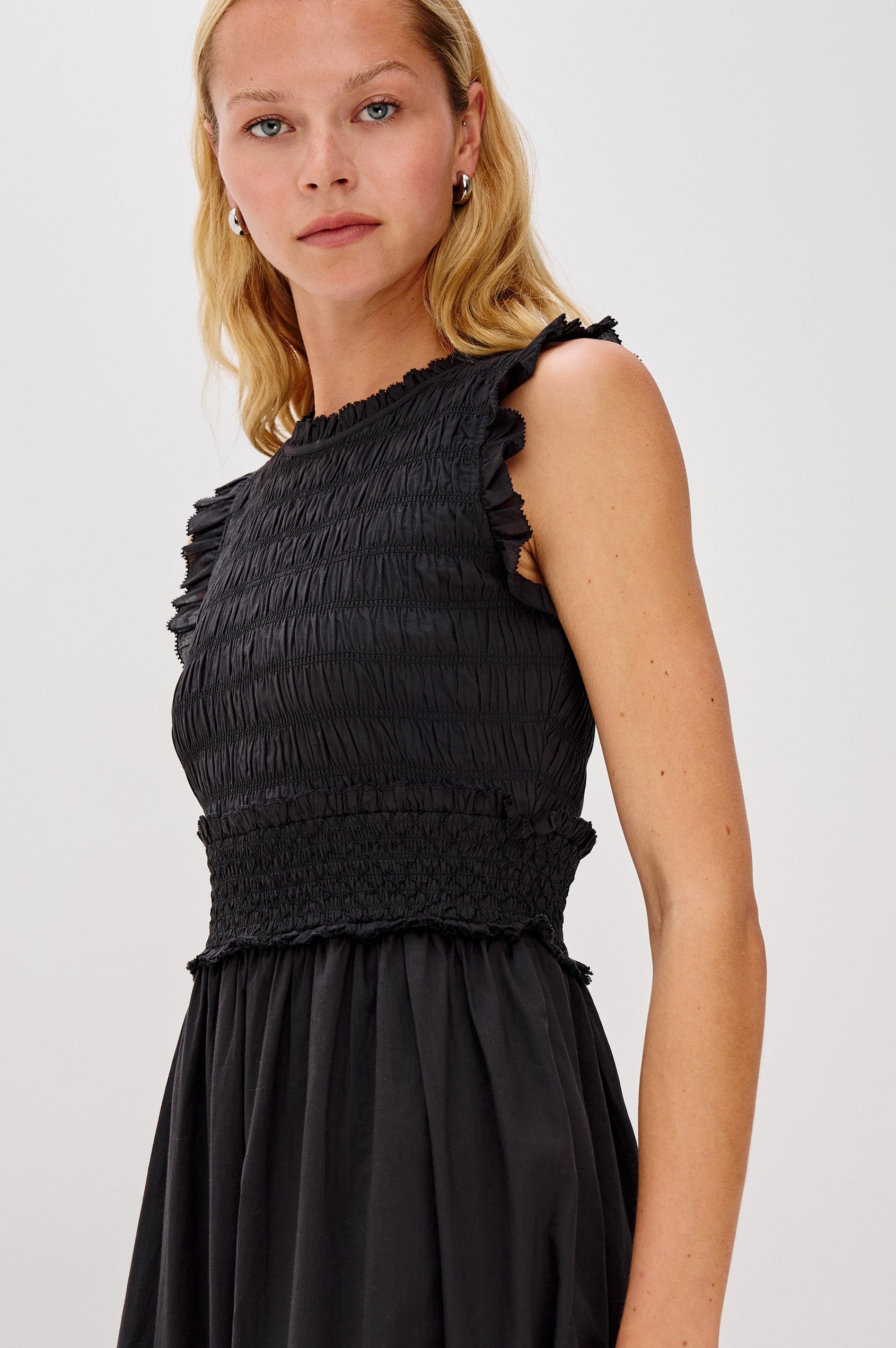 Vivier is a crinkled cotton poplin maxi dress featuring a smocked, fitted bodice and waistband. The dress flows into a relaxed skirt with soft ruffled tiers, creating a breezy silhouette in Black Colorway - Close up of upper Details on Model