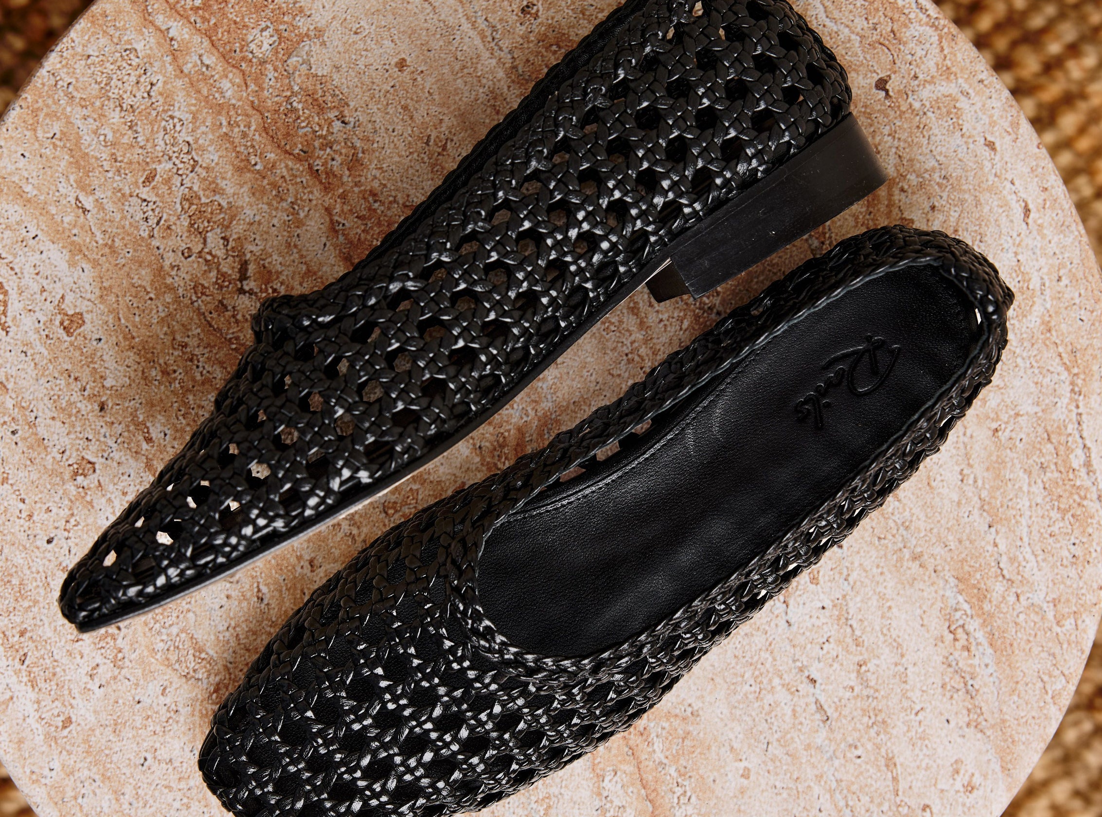 A pair of black pointed-toe flats with an open, woven-texture upper, displayed on a round beige stone slab.
