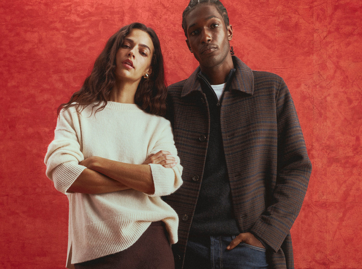 A man and a woman posing together against a textured red-orange background. The woman wears a cream-colored knit sweater, and the man wears a dark brown plaid button-up overshirt over a dark shirt and jeans.