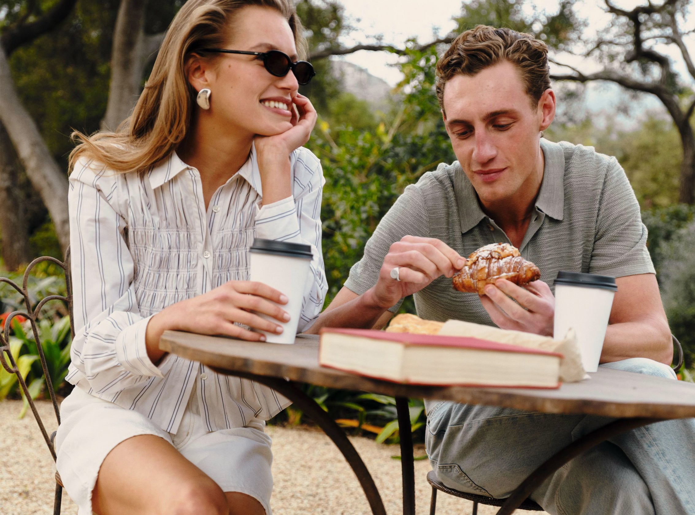 A man and a woman sitting at an outdoor cafe table, sharing coffee and a pastry. The woman is wearing a white button-down shirt with thin blue vertical stripes and white shorts, while the man wears a gray-green striped polo shirt and jeans.