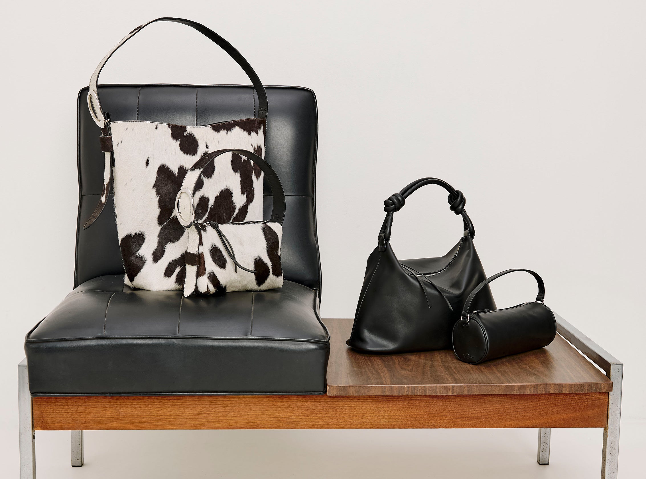 Two cow print handbags on black leather chair accompanied by two other black handbags on a table.