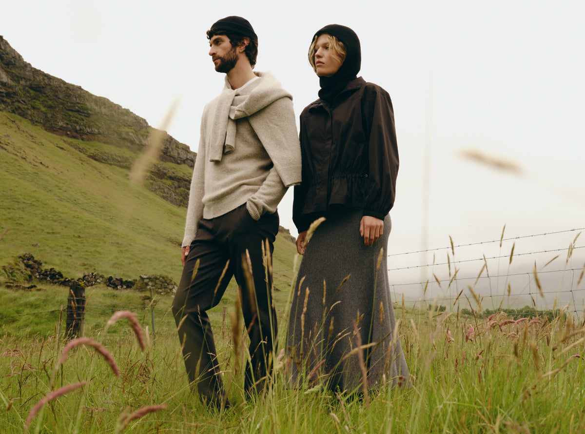 Two models standing in a grassy field with a hillside in the background, wearing earthy-toned outerwear. One wears a light beige knit sweater and dark trousers, and the other wears a dark brown hooded jacket and a long dark skirt.