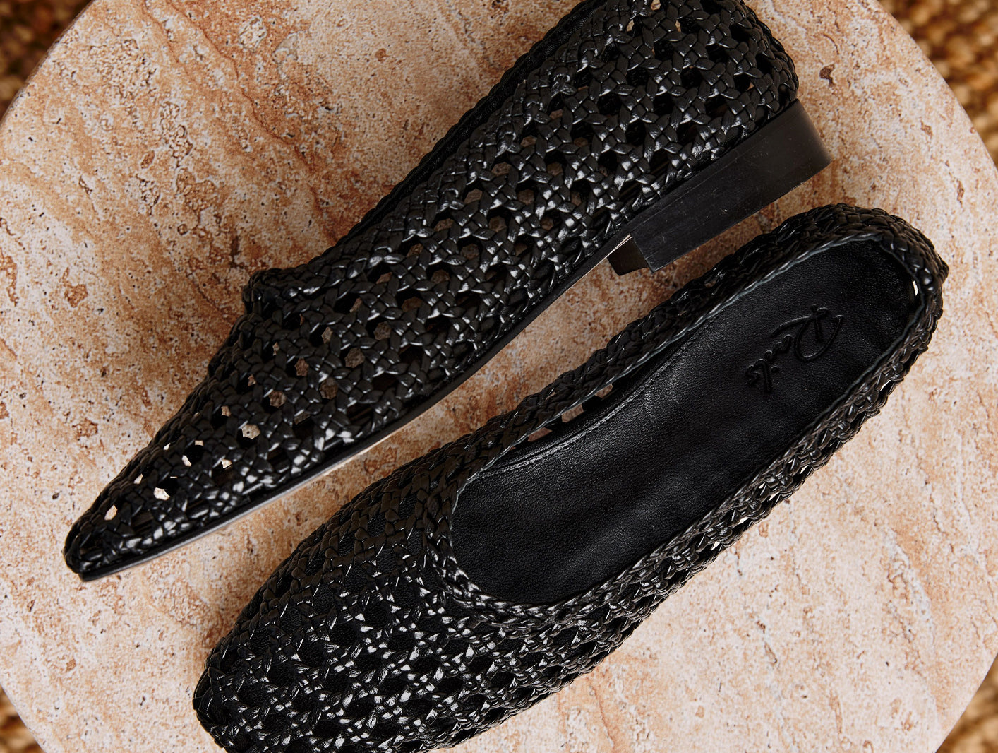 A pair of black pointed-toe flats with an open, woven-texture upper, displayed on a round beige stone slab.